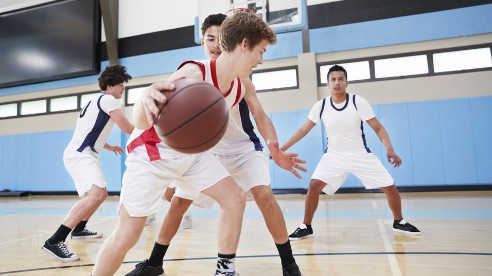 Vier Jugendliche spielen Basketball in einer Sporthalle, während ein Spieler den Ball dribbelt und von zwei Gegnern bewacht wird.