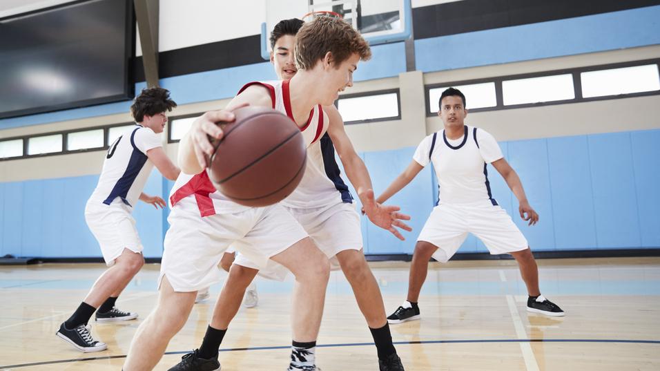 Vier Jugendliche spielen Basketball in einer Sporthalle, während ein Spieler den Ball dribbelt und von zwei Gegnern bewacht wird.