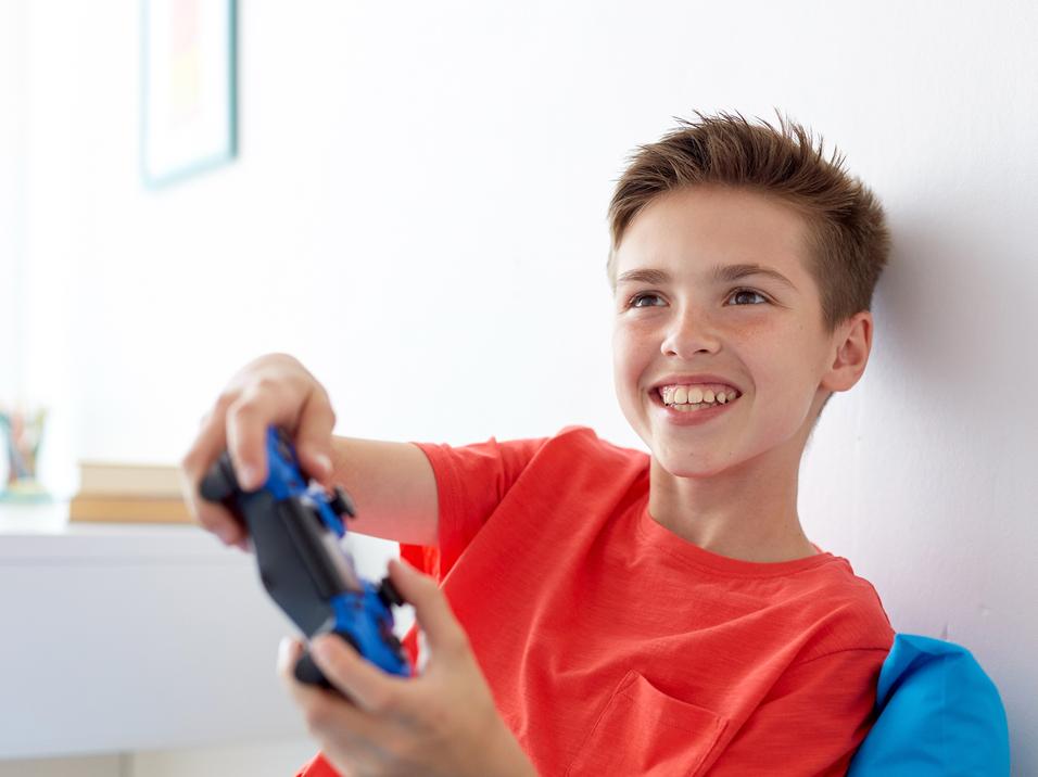 Junge sitzt auf einem blauen Kissen und spielt mit einem blauen Controller, lächelnd vor einer weißen Wand.