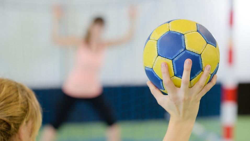 Nahaufnahme einer Hand, die einen gelb-blauen Handball hält, während eine Spielerin im Hintergrund positioniert ist.