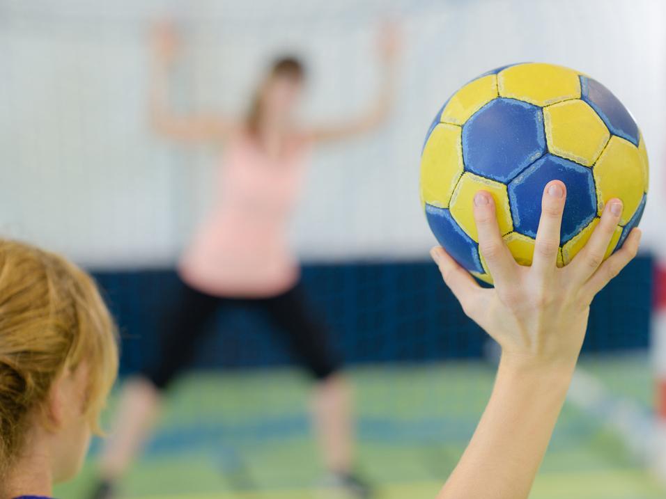 Nahaufnahme einer Hand, die einen gelb-blauen Handball hält, während eine Spielerin im Hintergrund positioniert ist.