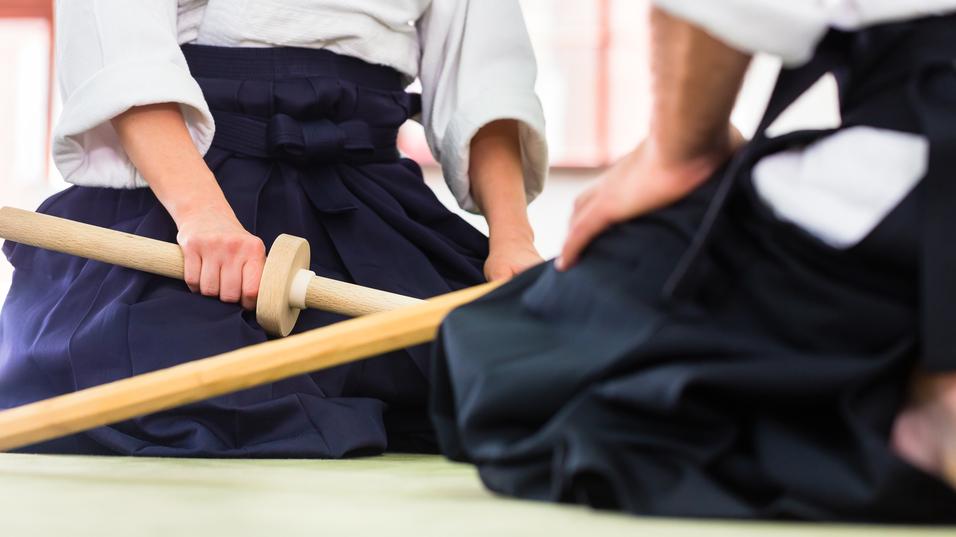 Zwei Personen in traditioneller japanischer Kleidung sitzen mit Holzschwertern auf einem grünen Boden.