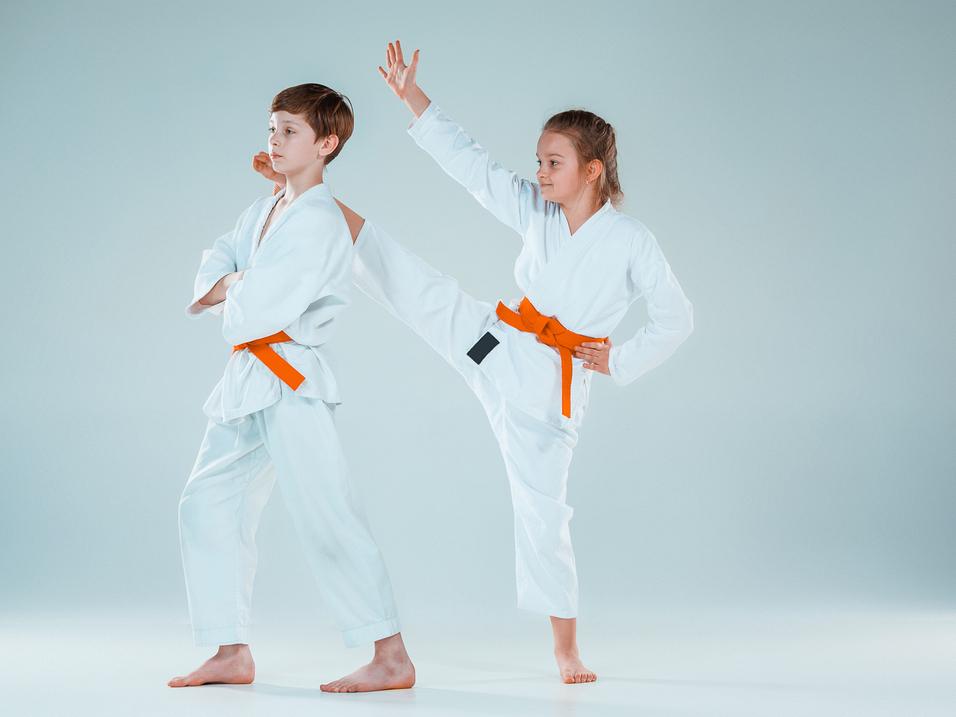 Zwei Kinder in weißen Karateanzügen mit orangefarbenen Gürteln, eines führt einen Kick aus, das andere steht still.