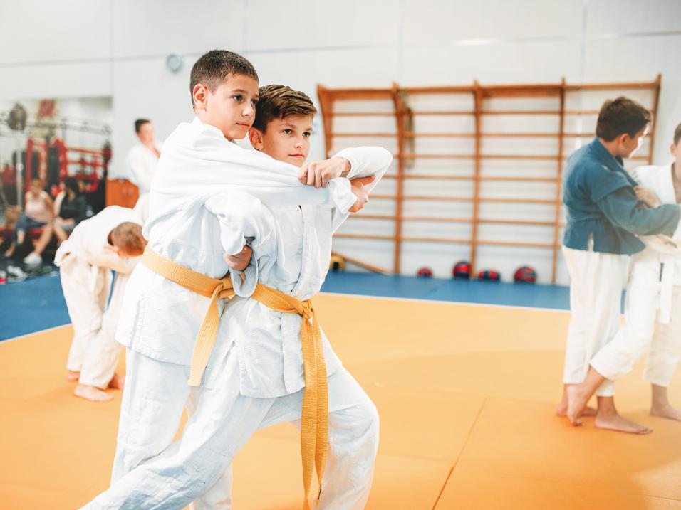 Zwei Jungen in weißen Judoanzügen üben eine Technik auf einer Judo-Matte, während andere im Hintergrund trainieren.