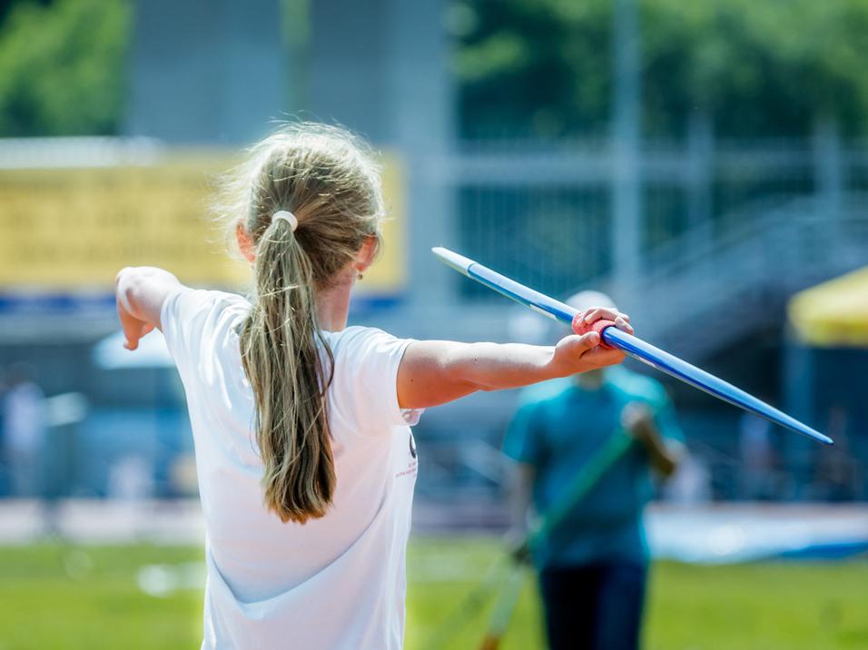 Mädchen wirft eine Sportspitze, trägt ein weißes T-Shirt und steht auf einer grünen Sportfläche.
