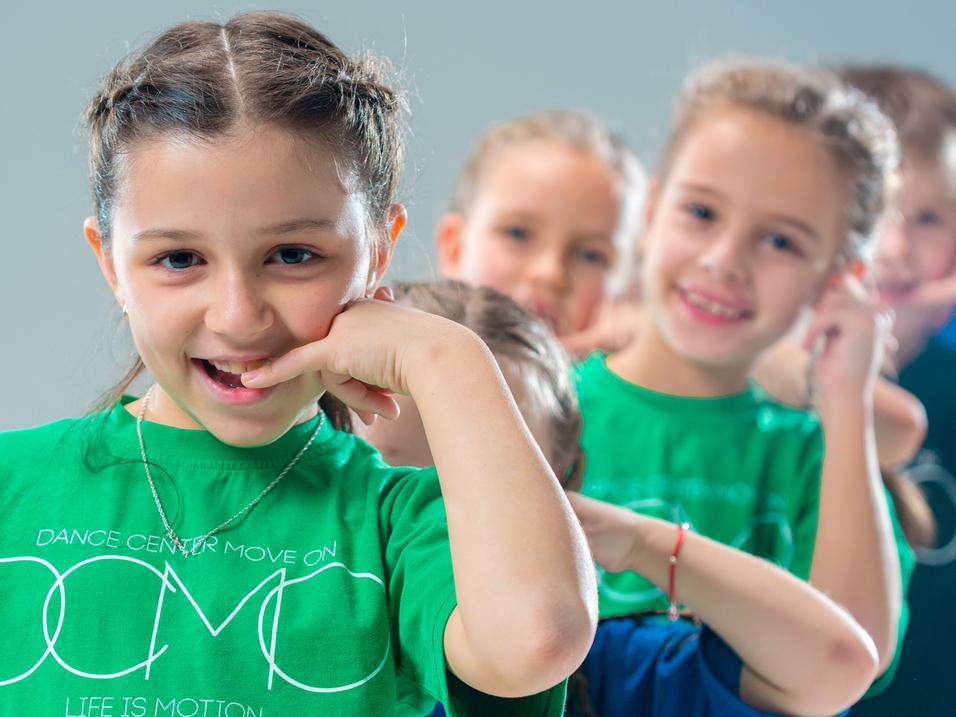 Lächelnde Kinder in bunten T-Shirts machen eine Tanzbewegung vor einem hellen Hintergrund.