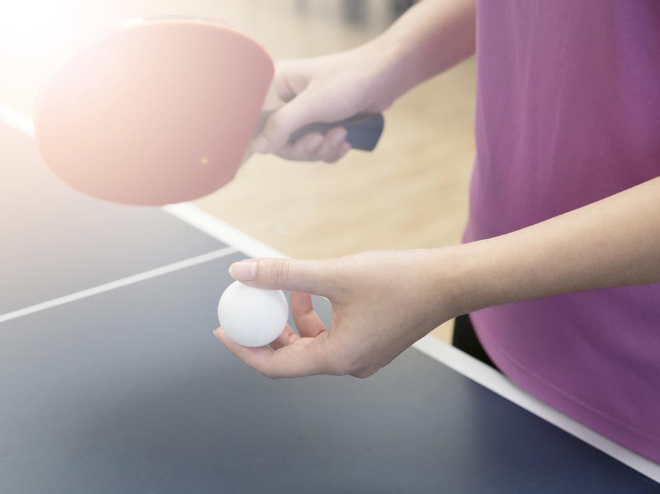 Eine Hand hält einen weißen Tischtennisball, während die andere einen Tischtennisschläger über einer Tischtennisplatte hält.