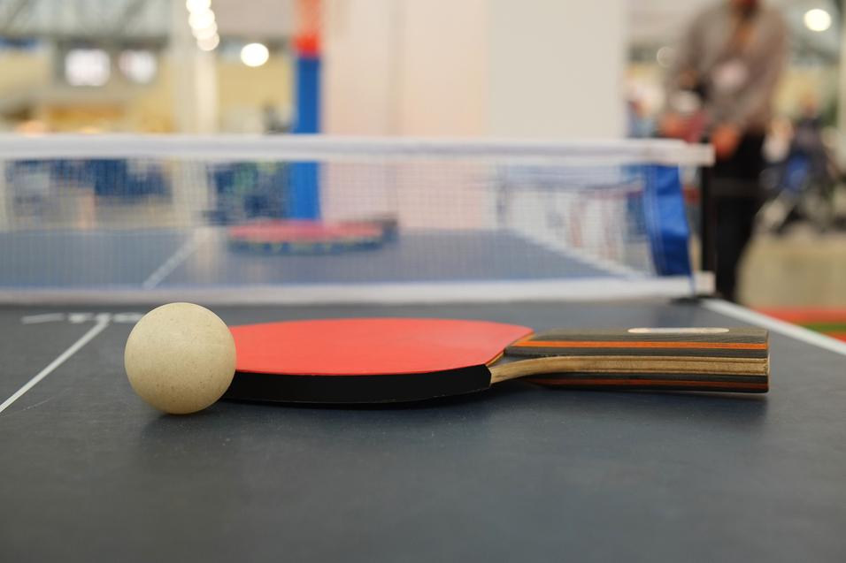 Nahaufnahme eines Tischtennisschlägers mit Ball auf einem Tischtennistisch mit Netz im Hintergrund.